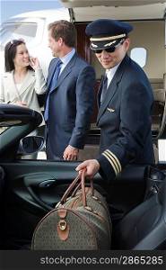 Mid-adult businesswoman and businessman standing in front of private jet, while pilot holding their luggage.