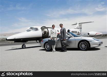 Mid-adult businesswoman and mid-adult businessman standing in front of convertible and private jet on landing strip.