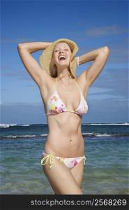 Mid-adult Caucasian female in bikini holding hat down with ocean in background.