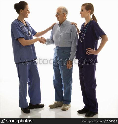 Mid-adult Caucasian female in scrubs shaking hand of elderly Caucasian male with another mid-adult Caucasian female with hand on his shoulder.