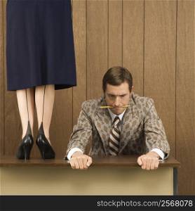 Mid-adult Caucasian male biting pencil and holding desk with Caucasian female standing on desk beside him.