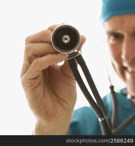 Mid-adult Caucasian male doctor in scrubs holding a stethoscope out towards viewer.