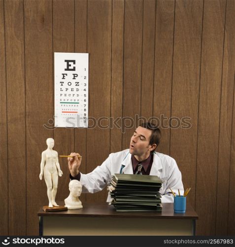 Mid-adult Caucasian male doctor sitting at desk pointing to figurine.