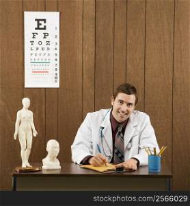Mid-adult Caucasian male doctor sitting at desk writing and looking at viewer.