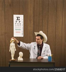 Mid-adult Caucasian male doctor wearing cowboy hat sitting at desk playing with figurine.