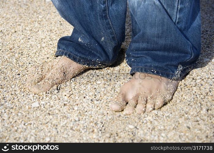 Mid-adult Caucasian male feet in jeans on sandy beach.