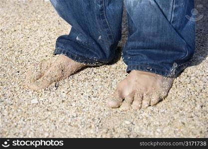 Mid-adult Caucasian male feet in jeans on sandy beach.