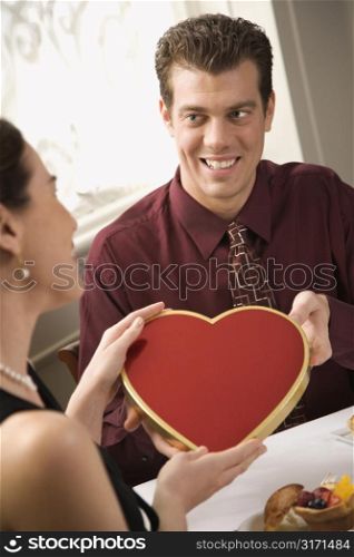 Mid adult Caucasian man giving a heart shaped box of chocolates to surprised woman at restaurant.