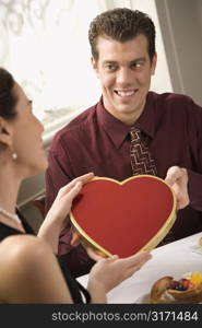 Mid adult Caucasian man giving a heart shaped box of chocolates to surprised woman at restaurant.