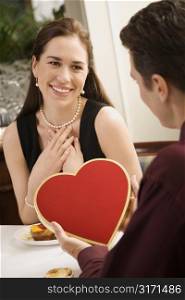 Mid adult Caucasian man giving a heart shaped box of chocolates to woman at restaurant.