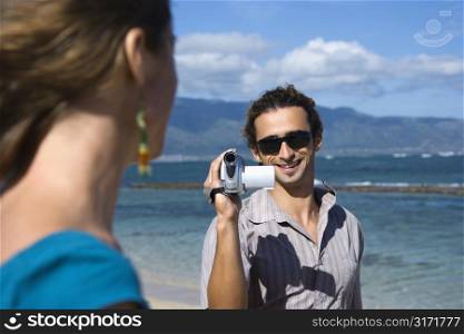 Mid-adult Caucasian man on beach pointing video camera at woman.