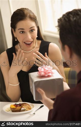 Mid adult Caucasian man presenting wrapped gift to surprised woman at restaurant.