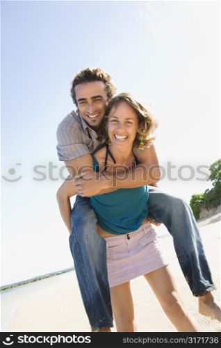 Mid-adult Caucasian woman giving man piggyback ride on beach.
