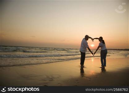 Mid-adult couple making heart shape with arms on beach at sunset.