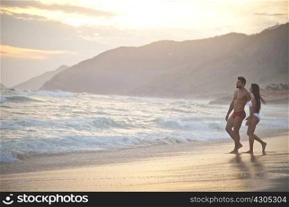 Mid adult couple on beach, wearing swimwear, walking towards ocean