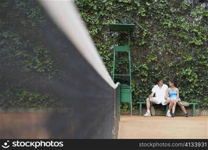 Mid adult man and a young woman sitting on a bench on a tennis court