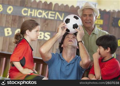 Mid adult man balancing a soccer ball on his forehead with his children and father looking at him