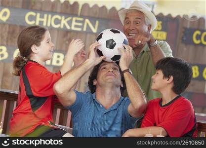 Mid adult man balancing a soccer ball on his forehead with his children and father looking at him