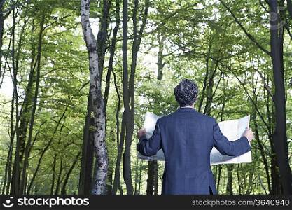 Mid adult man in suit jacket standing in forest with map, back view