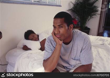 Mid adult man sitting on a bed with a mid adult woman lying down behind him