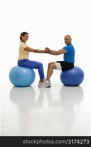 Mid adult multiethnic man and woman balancing on blue exercise balls with hands and feet locked together smiling and looking at viewer.