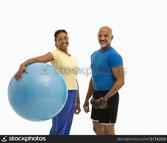 Mid adult multiethnic woman holding blue exercise ball standing with mid adult multiethnic man looking at viewer and smiling.