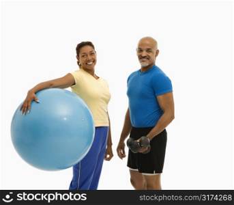 Mid adult multiethnic woman holding blue exercise ball standing with mid adult multiethnic man looking at viewer and smiling.
