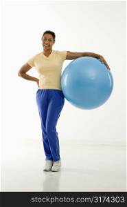 Mid adult multiethnic woman standing and holding blue exercise ball looking at viewer and smiling.
