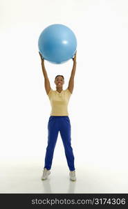 Mid adult multiethnic woman standing and holding blue exercise ball over her head looking at viewer and smiling.