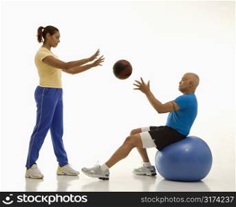 Mid adult multiethnic woman throwing ball to multiethnic mid adult man balancing on blue excersise ball.