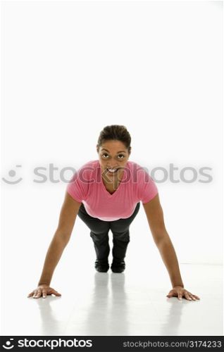 Mid adult multiethnic woman wearing pink exercise shirt doing pushups while looking at viewer and smiling.