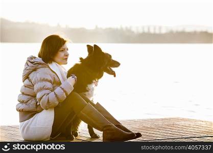 Mid adult woman and her dog sitting on sunlit lake pier