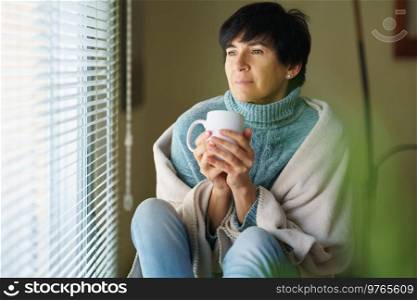 Mid adult woman drinking coffee and looking out of the window in winter day. Female in her 50s. Mid adult woman drinking coffee and looking out of the window in winter day.