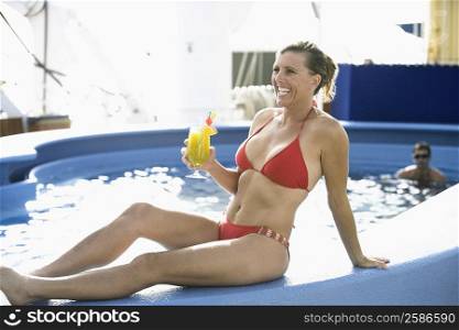 Mid adult woman holding a glass of juice and sitting on the ledge of a swimming pool