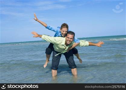 Mid adult woman riding piggyback on a mid adult man with their arms outstretched on the beach