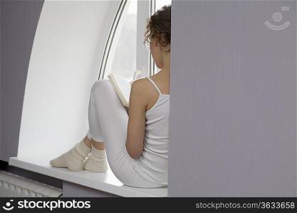 Mid adult woman sits on windowsill reading a book