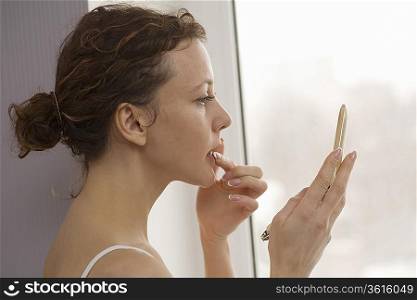 Mid adult woman stands applying lip gloss at window