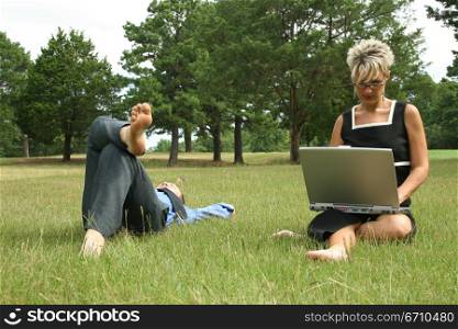 Mid adult woman working on a laptop with a man lying in the grass beside her