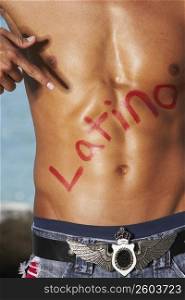 Mid section view of a bare chested young man pointing towards Latino written on his chest