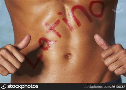 Mid section view of a bare chested young man pointing towards Latino written on his chest