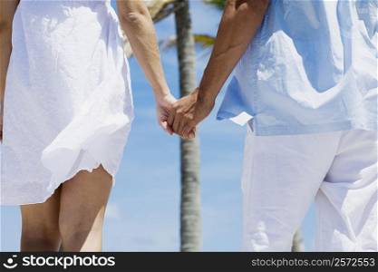 Mid section view of a couple holding hands of each other and walking on the beach