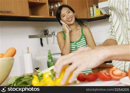 Mid section view of a man cutting vegetables with a young woman talking on a mobile phone in the kitchen