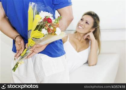 Mid section view of a man holding a bouquet of flowers behind his back with a young woman sitting on a couch and smiling