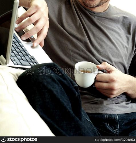 Mid section view of a man holding a cup of tea using a laptop