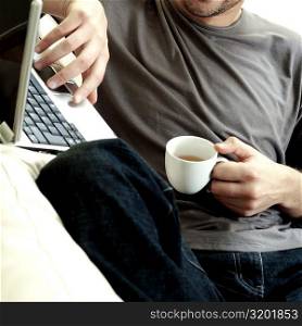 Mid section view of a man holding a cup of tea using a laptop