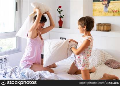 Middle-age mother with her eight years daughter having funny pillow fight on bed. Mum spending free time with her daughter.. Mother and daughter having funny pillow fight on bed.
