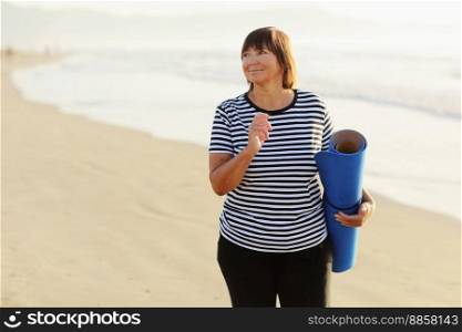 Middle age woman holding a sports mat and preparing to practice yoga outdoors on sea beach. Happy mature overweight woman exercising on seashore. copy space. Meditation, yoga and relaxation concept. Middle age woman holding a sports mat and preparing to practice yoga outdoors on sea beach. Happy mature overweight woman exercising on seashore. copy space. Meditation, yoga and relaxation concept.