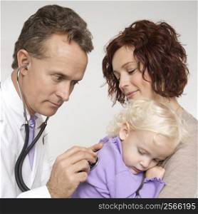Middle-aged adult Caucasian male doctor holding stethoscope to female toddler&acute;s back with mother watching.