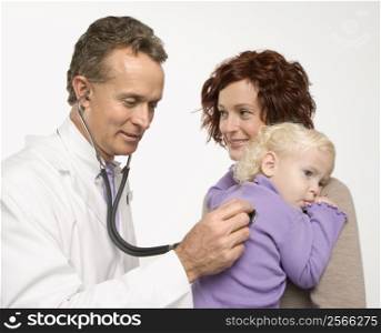 Middle-aged adult Caucasian male doctor holding stethoscope to female toddler&acute;s back with mother watching.