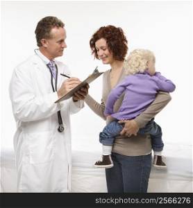 Middle-aged adult Caucasian male doctor taking notes on chart as mother holds daughter.